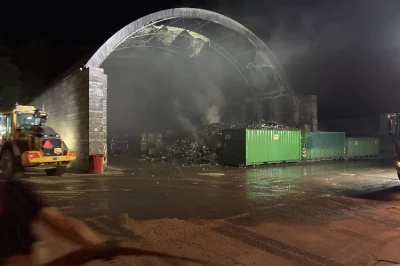 Rook stijgt op uit een brandende opslagruimte bij nacht, met een groene container op de voorgrond.