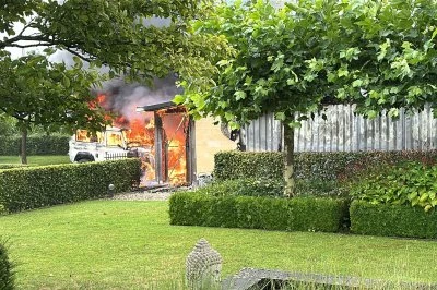 Brand in een schuur, omgeven door groen gras en struiken. Rook stijgt op.